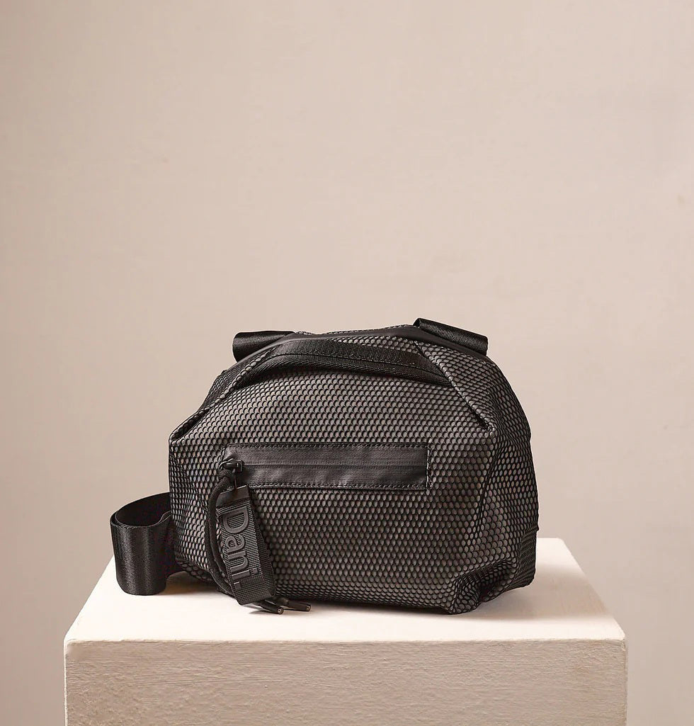 A person in a light beige suit carries the DANIELLA LEHAVI - BONNIE TRAVEL CROSSBODY, a large brown vegan bag with black zipper and strap, shown from shoulders to mid-thigh against a plain background.