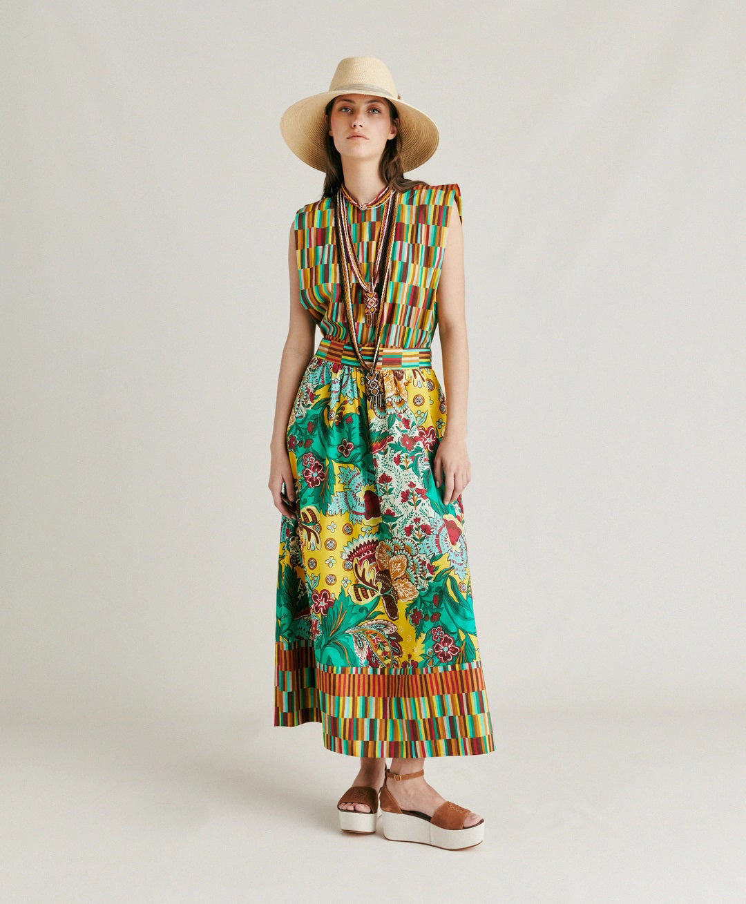 The MOMONI - IRIS SKIRT from MOMONI is a long, high-waisted skirt made from printed poplin. It features a vibrant floral and botanical design in green, yellow, red, and brown. The hemline and constructed waistband are accented with a contrasting striped pattern in similar hues. This skirt boasts a flowy, A-line silhouette.