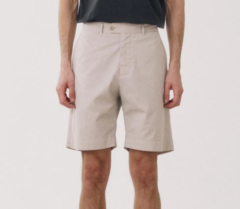 A man stands facing forward against a plain white background, wearing lightweight HARTFORD TAYLOR SHORTS in Stone, a dark short-sleeve T-shirt, and light sneakers.