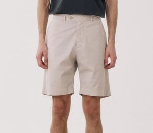 A man stands facing forward against a plain white background, wearing lightweight HARTFORD TAYLOR SHORTS in Stone, a dark short-sleeve T-shirt, and light sneakers.