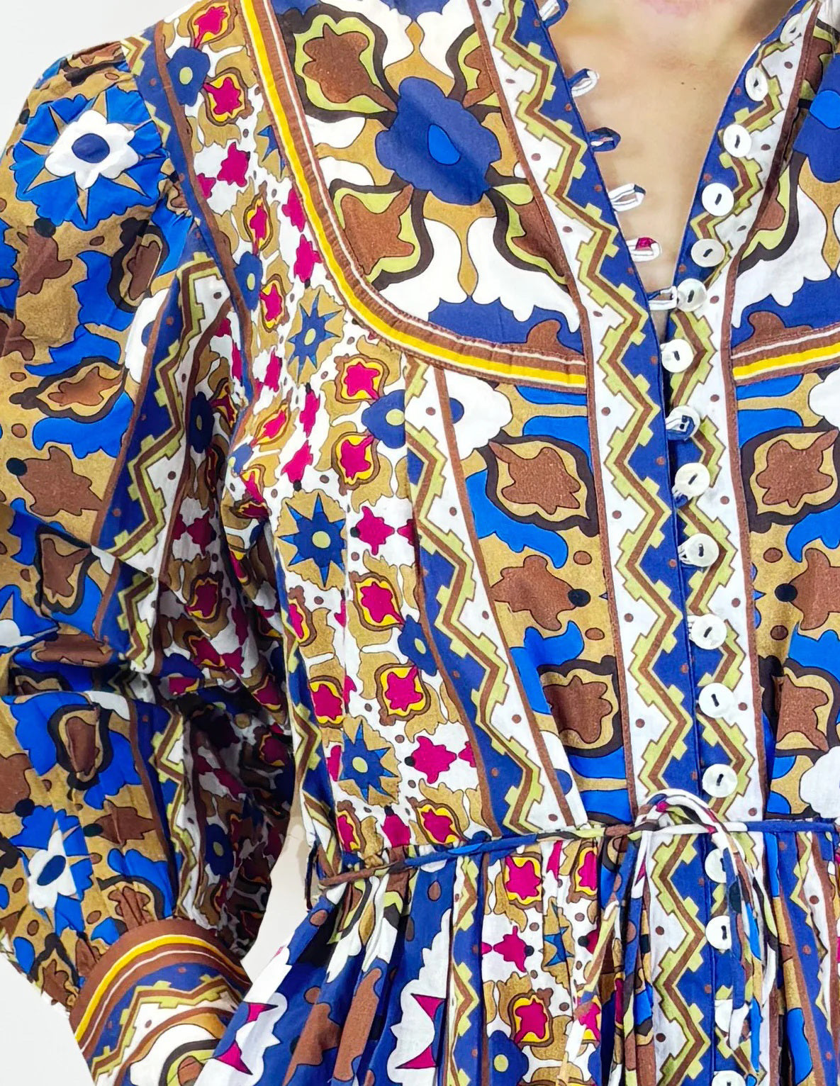 A woman poses against a plain background in the FARYLINA ANDI DRESS, a colorful bohemian style with long puff sleeves and a brown belt, styled with tan flats and long hair, one leg crossed in front of the other.