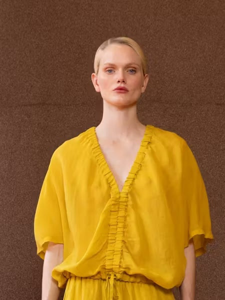 A person with light blonde hair in a low ponytail stands with their back to the camera, wearing the NUDE Tie Waist V Neck Blouse by NUDE in yellow, set against a plain brown background.