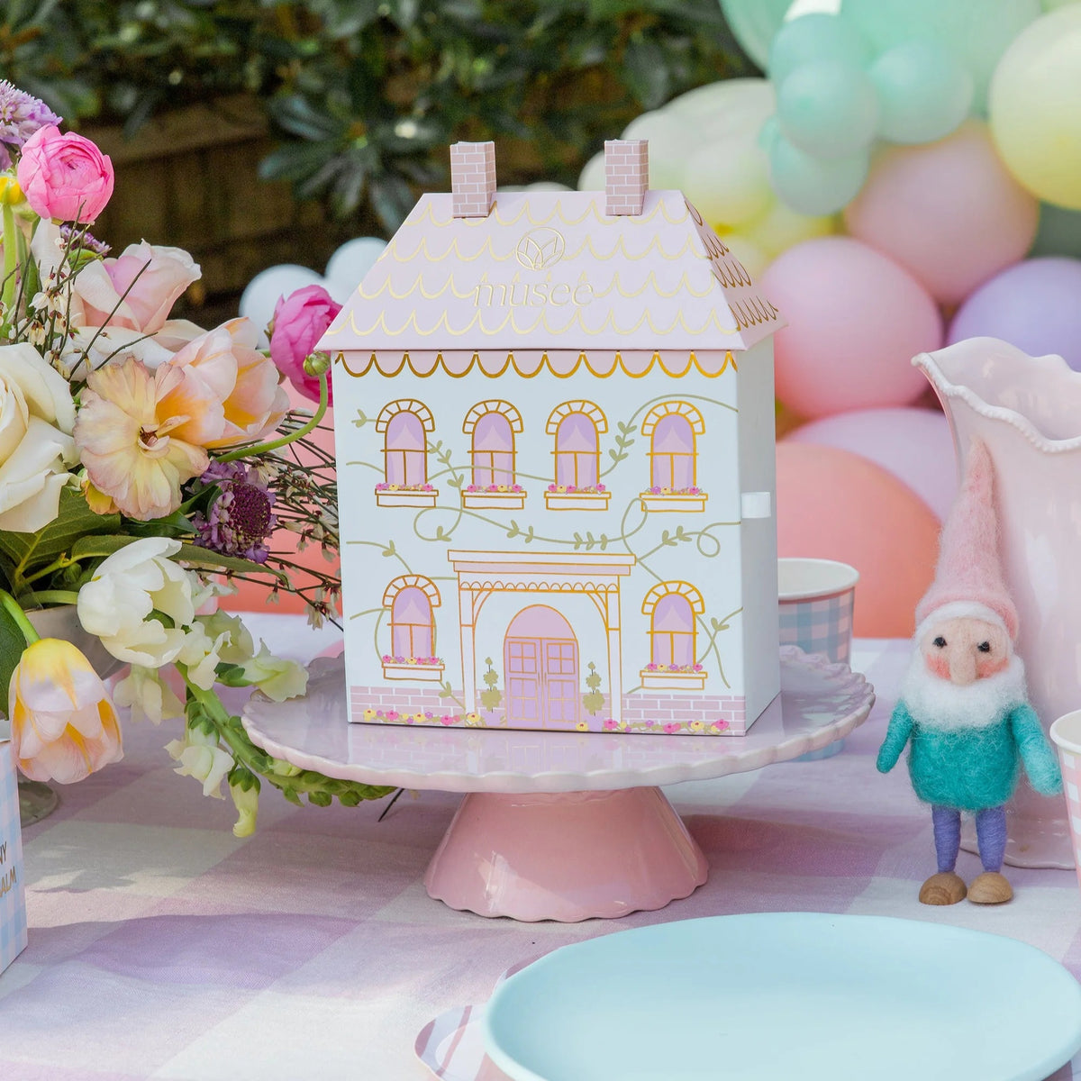 The MUSEE - DOLLHOUSE BATH BALM SET by MUSEE features a pastel, house-shaped gift box reminiscent of a dollhouse, complete with whimsical labeling. Inside, four naturally crafted bath balms—Fresh Linen, Flowering Lilies, Clean Citrus, and Warm Vanilla—are nestled in white decorative paper. This delightful set makes for a charming children’s gift.