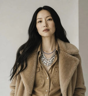A woman with long black hair wears a tan fur coat over a tan shirt and an ANN LIGHTFOOT - GREY PEARL FOUR STRAND FLIP FLOP COLLIER NECKLACE, standing against a plain light background.