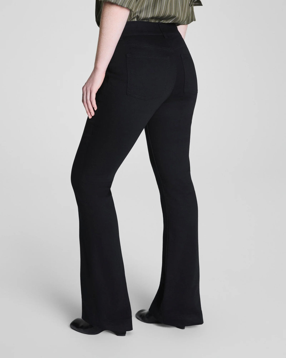 A person is shown from the back, wearing the SPANX - SPANXSHAPE EVERYWHERE PULL ON FLARE JEAN IN OBSIDIAN RINSE by SPANX with a green striped shirt, standing against a plain background to highlight the fit and style of the jeans.