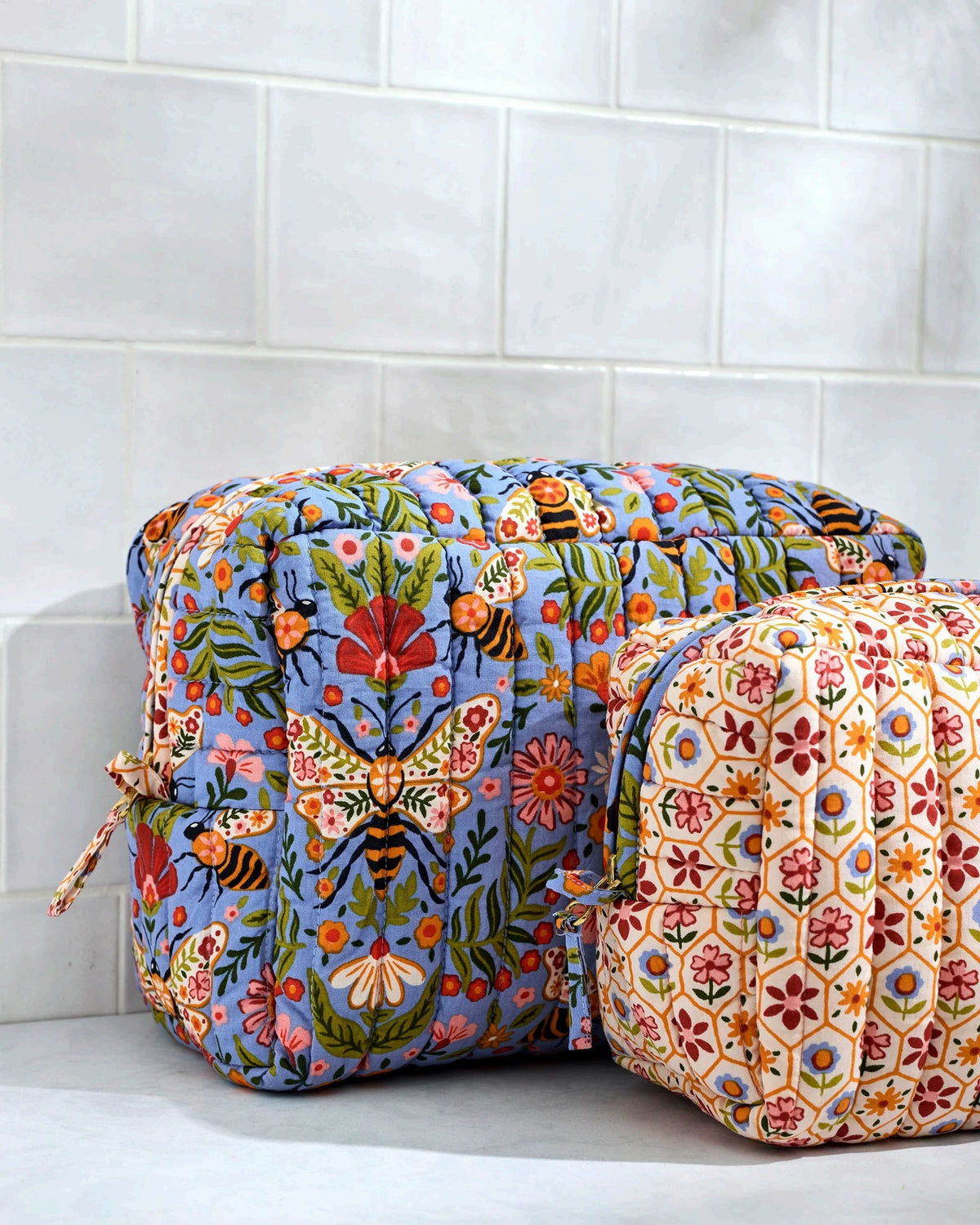 The PRINTFRESH Bee Tapestry Oversized Pouch Duo in Delphinium Blue features two quilted makeup pouches: the larger pouch showcases a blue bee and floral print, while the smaller pouch displays a cream background with red, yellow, and blue geometric shapes. Both PRINTFRESH bags are displayed on a tiled surface.