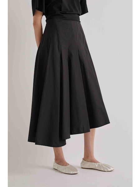 A person is seen from the back wearing a black short-sleeve top, the MEIMEIJ ASYMETRICAL COTTON PLEATED SKIRT, and white flat shoes, against a plain light background.