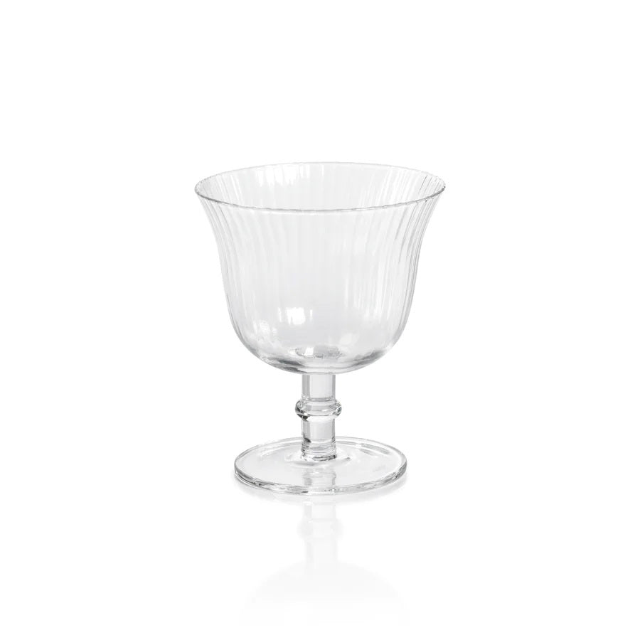 The JULIEN OPTIC COCKTAIL GLASS by ZODAX is a transparent glass goblet with a fluted pattern, featuring a wide bowl, a short stem, and a round base. With a capacity of 240 ml, the glass is empty and sits on a plain white background, creating reflections on the surface below.