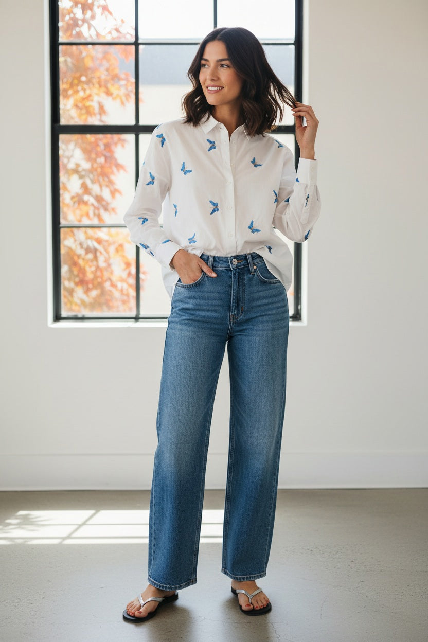 A woman with long, straight hair stands against a plain background wearing the 0039 ITALY LUCY BLOUSE WITH EMBROIDERED BUTTERFLIES, light blue wide-leg jeans, and silver sandals. She is looking slightly to the side.