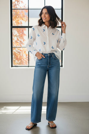 A woman with long, straight hair stands against a plain background wearing the 0039 ITALY LUCY BLOUSE WITH EMBROIDERED BUTTERFLIES, light blue wide-leg jeans, and silver sandals. She is looking slightly to the side.