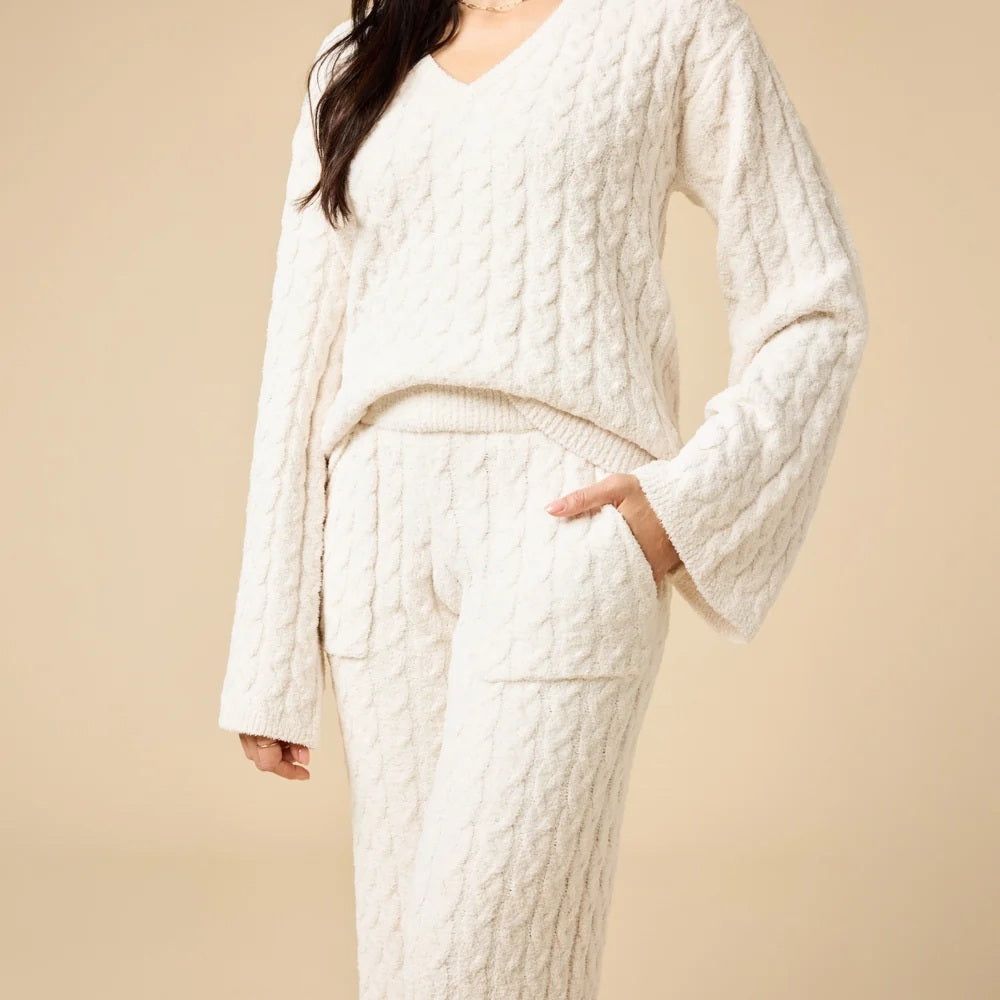 A woman wearing the SOFTIES CABLE KNIT MARSHMALLOW V-NECK WITH FLARED SLEEVE AND PANT SET, a marshmallow-soft, cream-colored cable knit v-neck sweater with wide sleeves and matching lounge pants.
