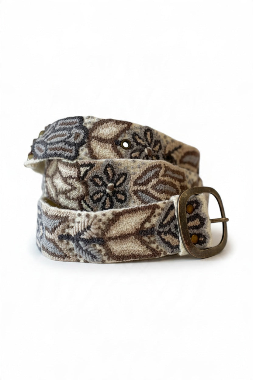 The JENNY KRAUSS Monochromatic Belt in Cream, handcrafted in Peru with floral and geometric embroidery in beige, brown, and gray, features a metal buckle and is shown coiled on a light surface.