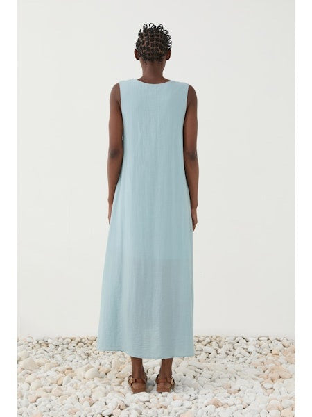 A person stands on white pebbles wearing the ELEH - DRESS WITH CENTRAL KNOT, a sleeveless sky blue A-line dress by ELEH, paired with brown sandals, against a plain white background.
