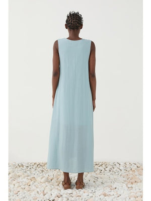 A person stands on white pebbles wearing the ELEH - DRESS WITH CENTRAL KNOT, a sleeveless sky blue A-line dress by ELEH, paired with brown sandals, against a plain white background.
