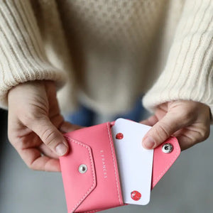 A person in a cream sweater holds the E. FRANCES - E. FRANNIE PACK, BUBBLEGUM wallet by E. FRANCES PAPER, pulling out a white card with red hearts—an adorable gift for spreading joy.