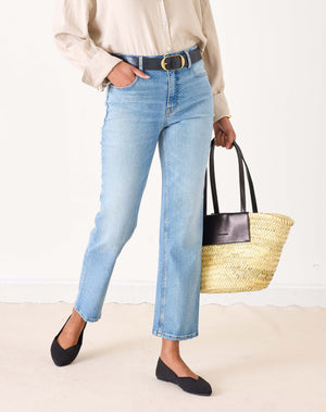 A person wearing a beige button-up shirt, MERSEA Bessie Straight Leg Jean in Caribbean made from recycled cotton, and black flats stands holding a woven straw tote bag with black handles. Their left hand is in their pocket, face not visible.