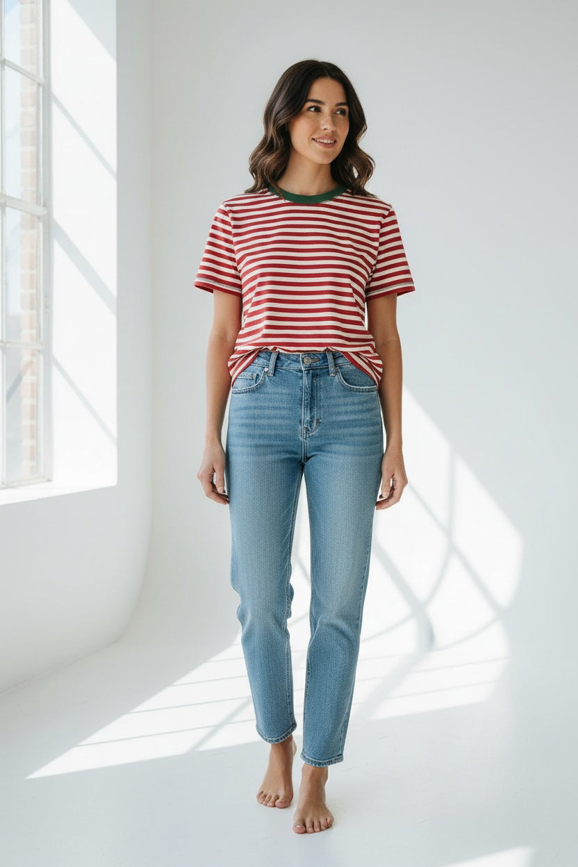 A smiling person with long blonde hair wears the MERSEA Deauville Ringer Tee in Currant Jade—a red and white striped ringer tee with green collar and cuffs—paired with green pants, standing against a light background.