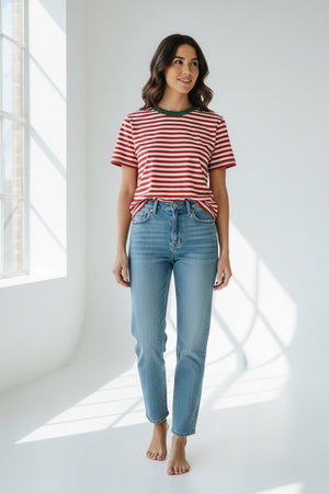 A smiling person with long blonde hair wears the MERSEA Deauville Ringer Tee in Currant Jade—a red and white striped ringer tee with green collar and cuffs—paired with green pants, standing against a light background.