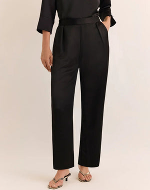 A person is wearing HINSON WU’s HOSTESS LIQUID SATIN PANT IN BLACK—high-waisted, wide-leg pants crafted from lustrous liquid satin fabric—paired with a black three-quarter sleeve top and open-toe heels with a thin strap. Their left hand is in the pocket of the pants, and their head is not visible.