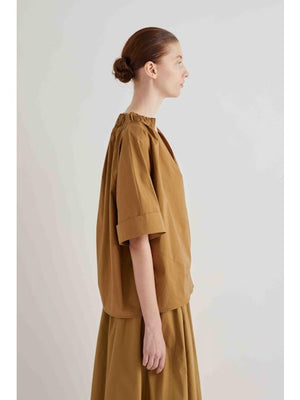 A woman with her hair in a bun stands in profile, wearing the MEIMEIJ Gathered Neck Blouse in Black by MEIMEIJ against a plain light background.