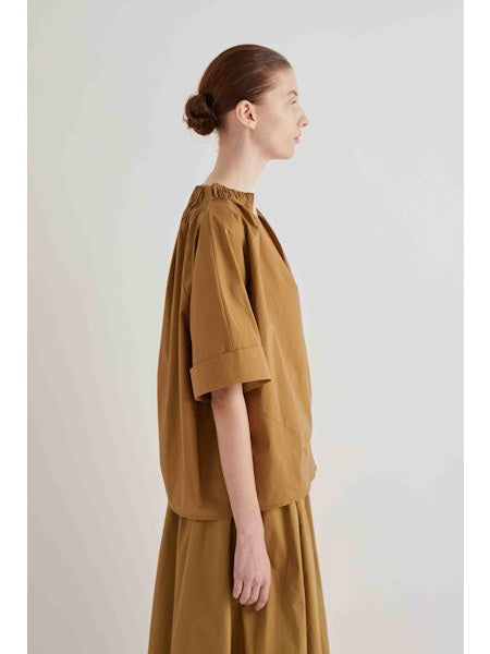 A woman stands against a plain background wearing the MEIMEIJ Gathered Neck Blouse in oak with short sleeves, paired with a matching midi skirt featuring a cut-out floral hem and black sandals.