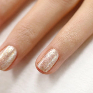 A hand with medium-brown skin displays short, neatly manicured nails painted with MANUCURIST - GREEN™ NAIL POLISH IN GOLD by MANUCURIST INC, featuring a shimmery pale gold glitter finish against a neutral beige background.