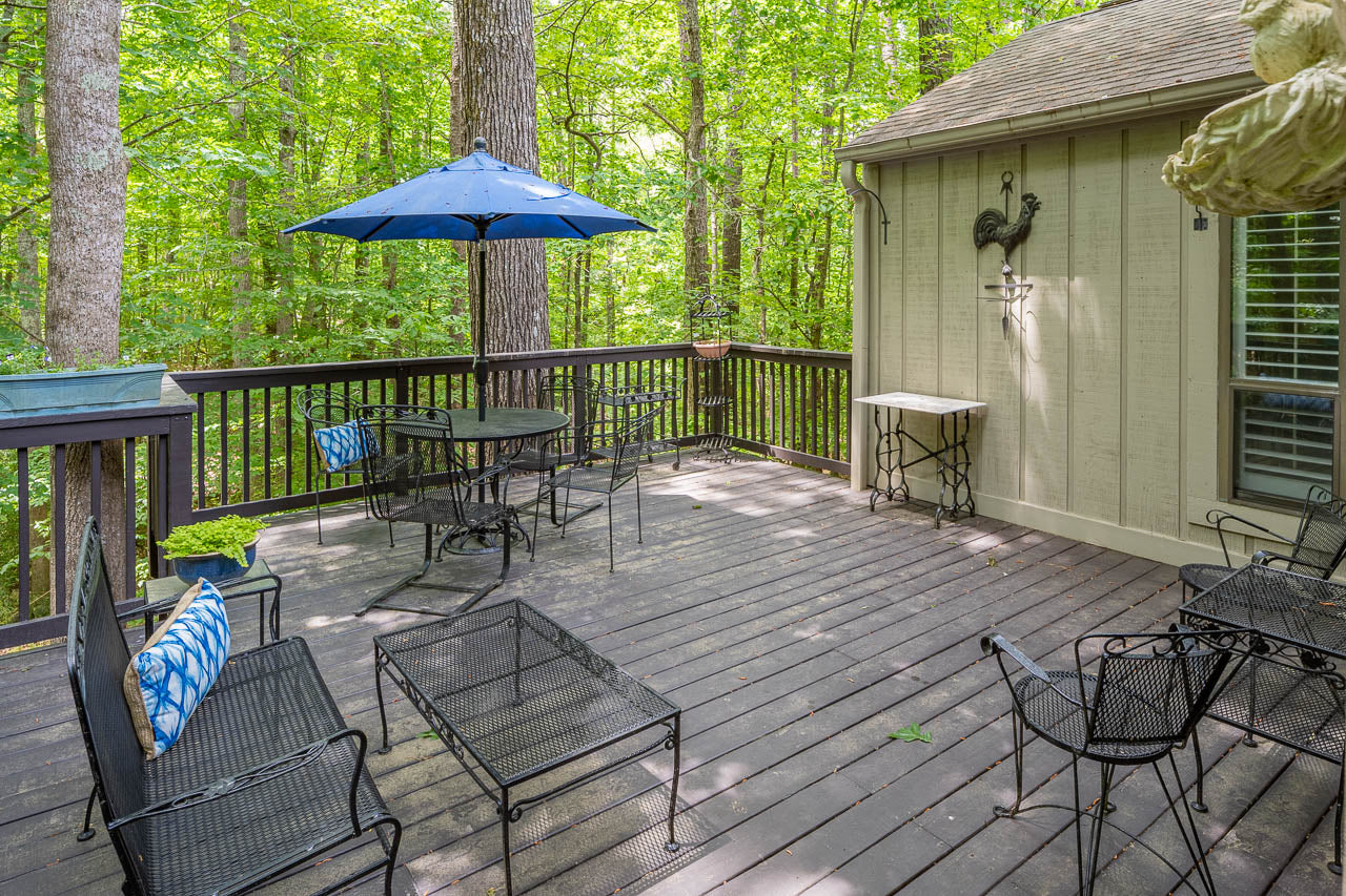 A wooden deck with black metal patio furniture, a blue umbrella, and a decorative wall piece, surrounded by lush green trees and shaded by the forest.