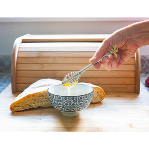 A hand holds an ABBOTT BEE HANDLE HONEY DAUBER, drizzling honey into a patterned bowl next to bread.
