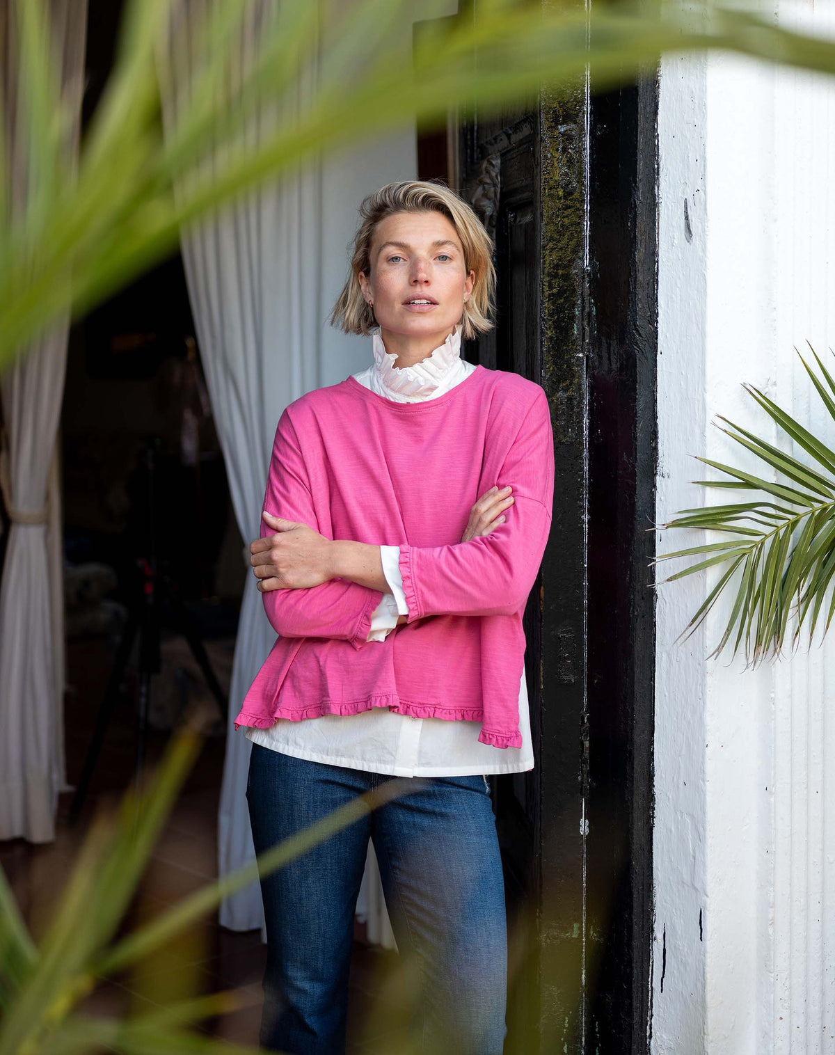 A person with short blonde hair stands in a doorway, arms crossed, wearing the MERSEA Catalina Ruffle Tee in Rapture Rose layered over a white blouse with blue jeans. Green leaves frame the foreground.
