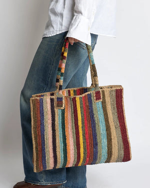 The GABRIELE FRANTZEN LITTLE SISTER WEEKENDER BAG CIRCUS is a large rectangular raffia bag with colorful vertical stripes, two matching handles, and a flat base—perfect as a stylish weekender.