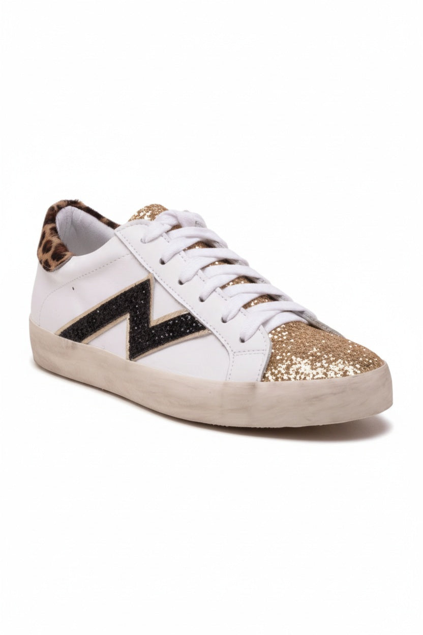 The MICHELE LOPRIORE - NINA SNEAKER features soft white leather, a black heel, glittery silver toe cap, zigzag black and beige side detailing, and a durable rubber sole, presented on a white background.