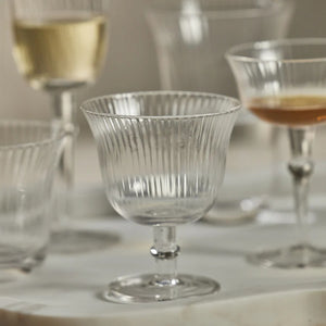 The JULIEN OPTIC COCKTAIL GLASS by ZODAX is a transparent glass goblet with a fluted pattern, featuring a wide bowl, a short stem, and a round base. With a capacity of 240 ml, the glass is empty and sits on a plain white background, creating reflections on the surface below.