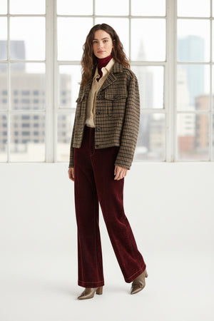 A woman stands indoors in front of tall windows, wearing a plaid jacket, a cream turtleneck, and the MOMONI EDIMBURGO Pant in Burgundy by MOMONI—flair trousers with a low-rise waist—paired with brown heeled shoes. Urban buildings are visible outside.