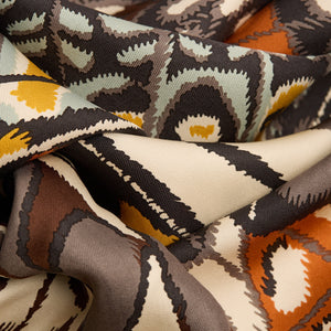 A close-up of a person wearing the INOUI EDITIONS - PAPILLON (BUTTERFLIES) SQUARE WOOL SCARF IN NATURAL by INOUI, draped around their neck over a black top. The scarf showcases abstract and geometric butterfly designs in earthy tones.