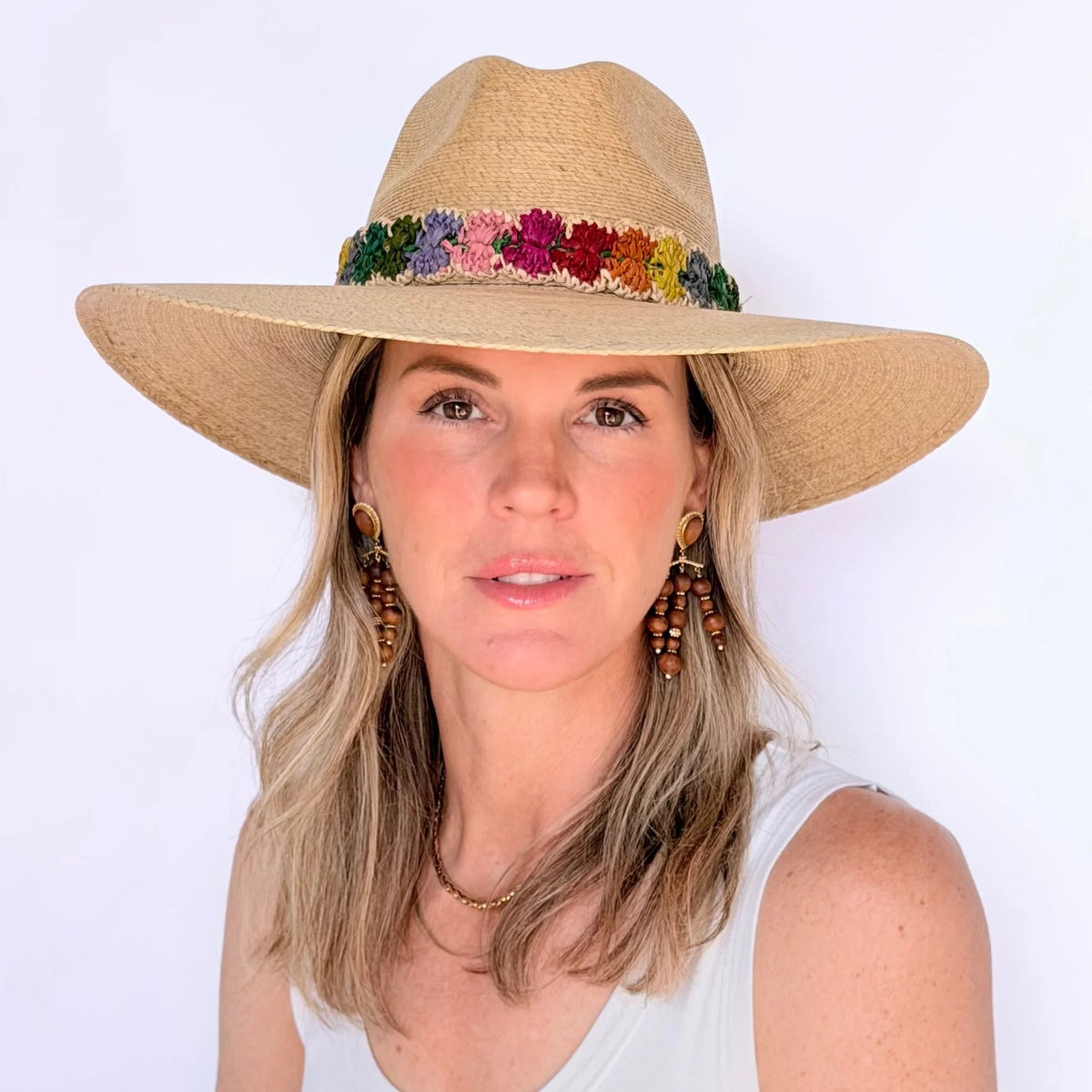 A woman with blonde hair models the BALDIZ Sand Palm Hat with Rainbow Band, pairing it with wooden earrings and a white sleeveless top against a plain white background. This stylish one-size-fits-most hat is by BALDIZ.