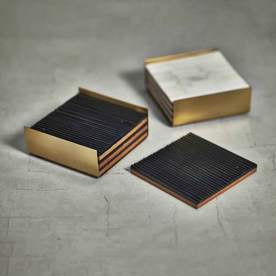 A set of RIBBED BONE COASTERS S4 by ZODAX, highlighting black-striped coasters neatly arranged in a gold metallic holder, with one coaster placed outside. The 4 in x 4 in coasters and holder showcase a modern, sleek design and are displayed on a white surface.