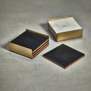 A set of RIBBED BONE COASTERS S4 by ZODAX, highlighting black-striped coasters neatly arranged in a gold metallic holder, with one coaster placed outside. The 4 in x 4 in coasters and holder showcase a modern, sleek design and are displayed on a white surface.