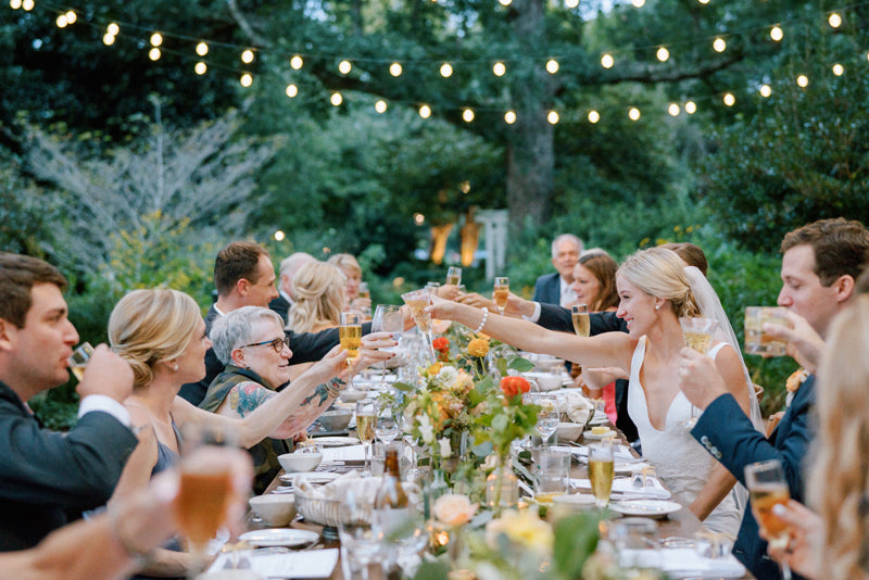 Wedding guests sit at a long, outdoor table decorated with flowers and string lights, raising glasses in a celebratory toast. The bride and groom sit together, smiling and toasting with others. Lush greenery surrounds the festive scene.