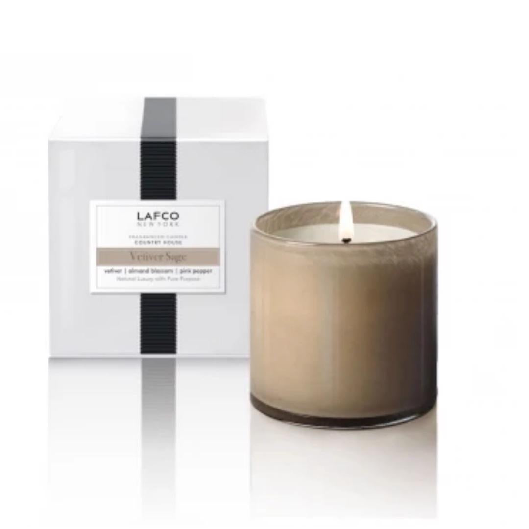 A lit LAFCO - VETIVER SAGE, COUNTRY HOUSE candle in a gray glass holder sits next to its white and black packaging box, which is labeled with &quot;LAFCO&quot; and the scent name &quot;Vetiver Sage.&quot; This hand-poured room candle emits a warm, soothing glow that is ideal for adding a touch of elegance to any country house.