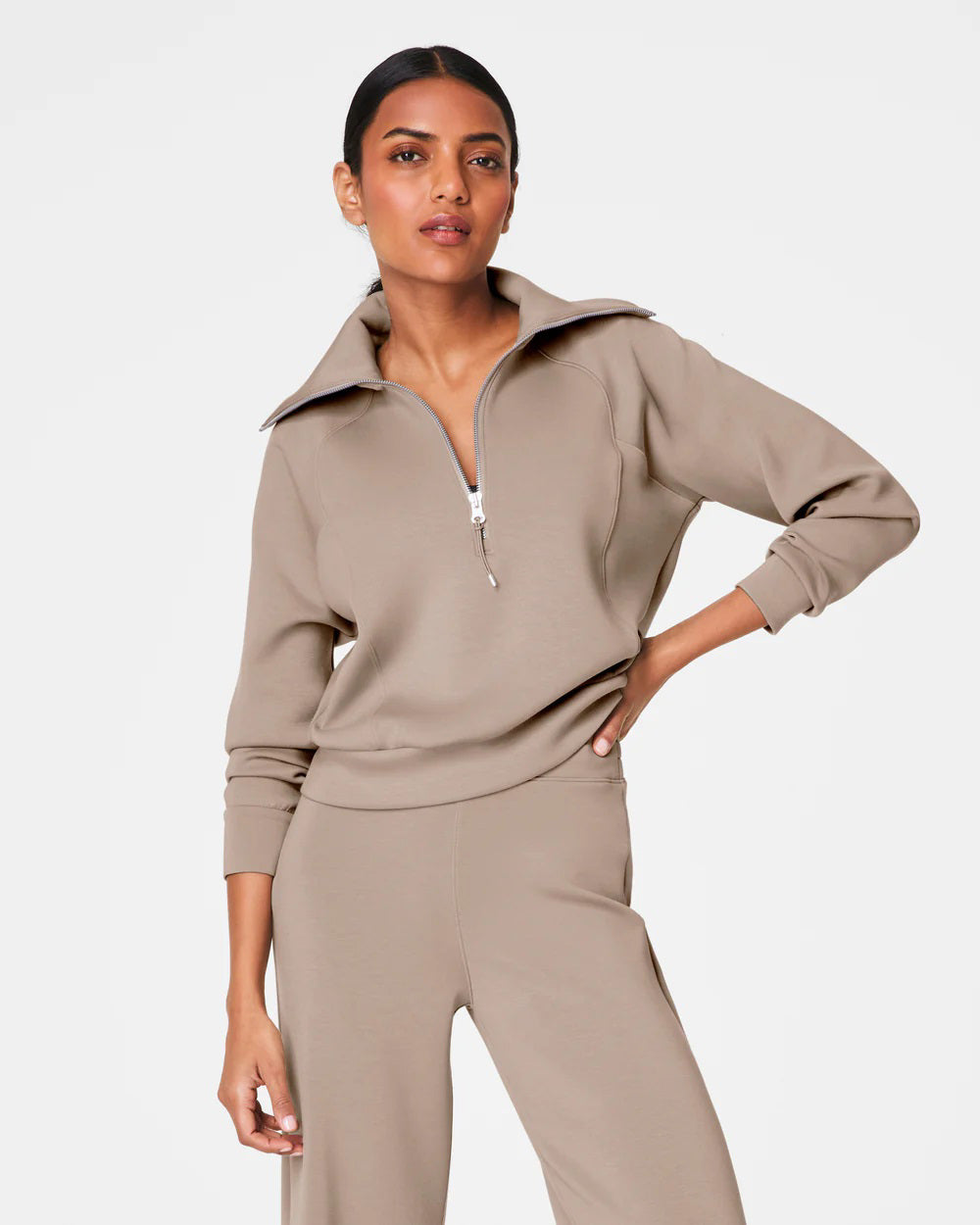 A person with short hair is wearing a SPANX AIRESSENTIALS HALF ZIP sweater in navy blue, made from lightweight spacer fabric, paired with black pants. Their arms are crossed over their chest as they stand against a plain, light grey background.