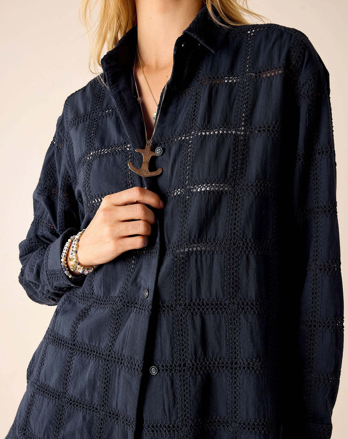 A woman with long blonde hair wears the MERSEA Brontë Button Up Shirt in Navy Crochet—a relaxed, textured long-sleeve shirt—paired with matching shorts, standing in profile against a light beige background.
