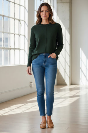 A woman with long, light brown hair wears the KINROSS CASHMERE - HI LOW SEAMED CREW NECK CASHMERE SWEATER IN HIGHLAND and blue jeans. She stands facing forward against a plain white background, smiling slightly.
