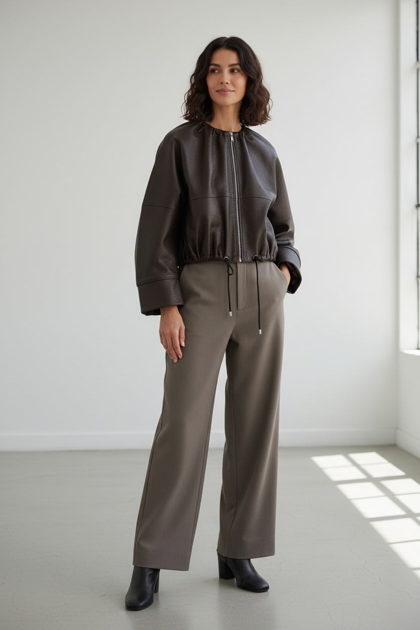 A person wearing the MEIMEIJ FAUX LEATHER BOMBER JACKET in dark brown faux leather with a textured finish and adjustable drawstring hem, paired with taupe high-waisted pants, stands against a plain light background. The jacket by MEIMEIJ lends a modern edge to the outfit, while the person’s head is partially out of frame.