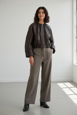 A person wearing the MEIMEIJ FAUX LEATHER BOMBER JACKET in dark brown faux leather with a textured finish and adjustable drawstring hem, paired with taupe high-waisted pants, stands against a plain light background. The jacket by MEIMEIJ lends a modern edge to the outfit, while the person’s head is partially out of frame.