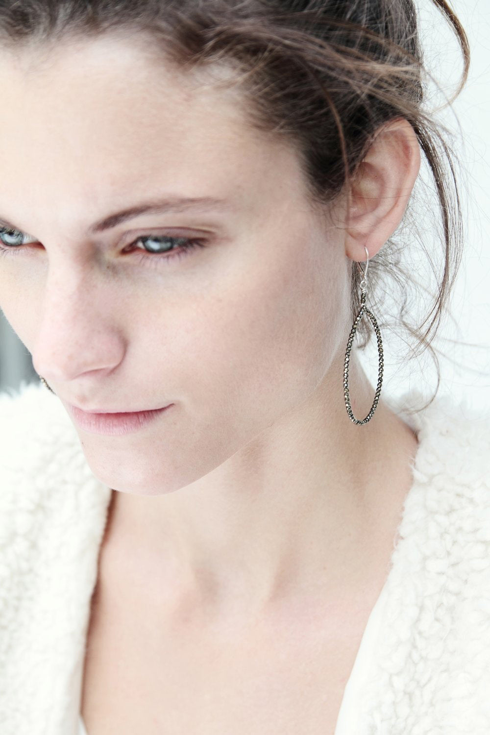 A light-skinned woman with a subtle expression gazes downward, dark hair pulled back and adorned with ANN LIGHTFOOT&#39;s Faceted Pyrite Hoop Earrings. Her soft-textured clothing contrasts against a bright, softly focused background.