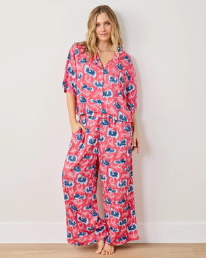 Against a neutral wall, a person stands barefoot in PRINTFRESH’s Satin Ostrich Dance Wildest Dream Pajama Set. The bright red ensemble with blue and white floral patterns features a short-sleeved button-up top and wide-legged pants, exuding comfort and style as they smile warmly.