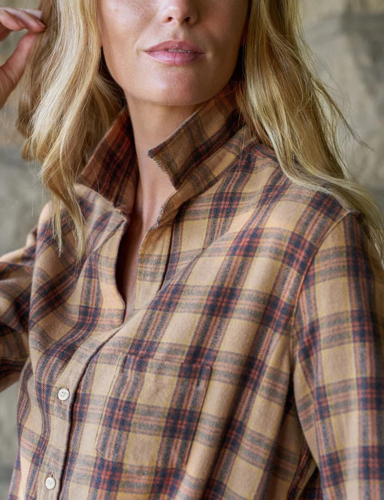 A woman stands in a sunlit room, smiling with curly hair, wearing the FRANK & EILEEN EILEEN Relaxed Button Up Shirt in Italian Flannel Gold Brown Plaid, paired with blue skinny jeans and brown ankle boots.
