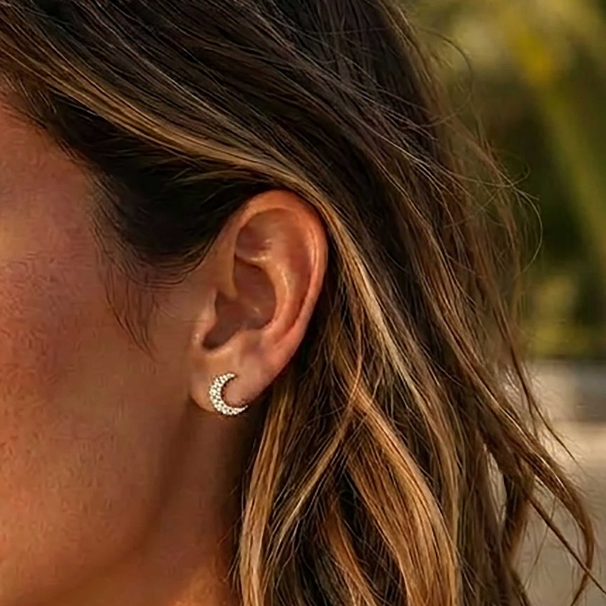 A close-up of a woman’s ear wearing the ELLA STEIN - MINI MOON STUD EARRINGS by ELLA STEIN, featuring small crescent moon shapes adorned with sparkling natural diamonds set in gold-plated stainless steel. Brown hair with blonde highlights is visible in the background.
