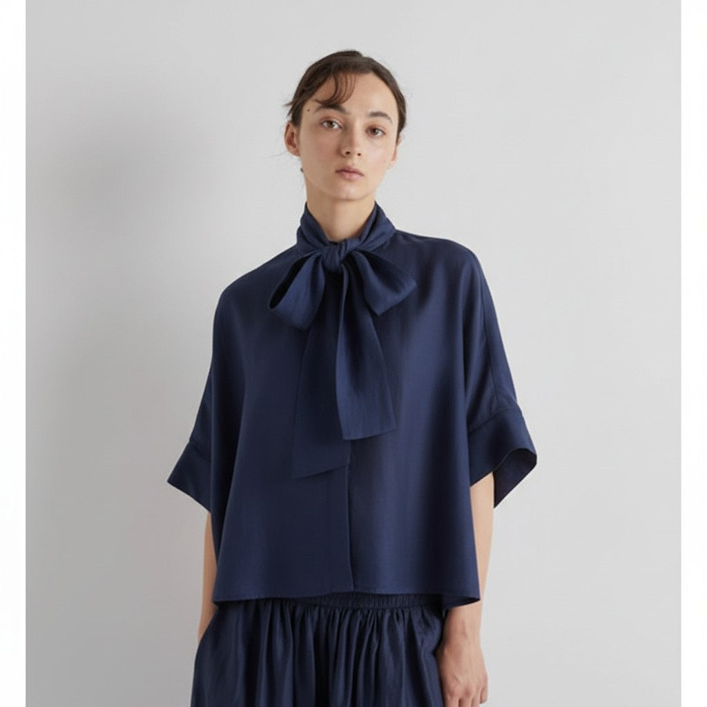 A woman models the MEIMEIJ SCARF TIE NECK BLOUSE in navy, featuring short sleeves and a bow tie neck, paired with a matching skirt, against a plain light gray background.