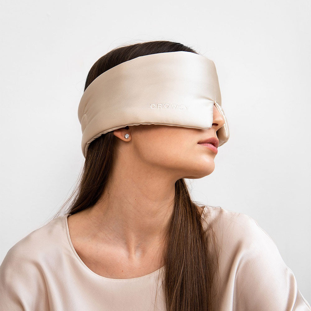 An older person with long gray hair wears a luxurious beige DROWSY - SATIN SLEEP MASK by DROWSY SLEEP CO, made of mulberry silk. In a matching satin top, they stand against a plain white background, ready for beauty sleep.